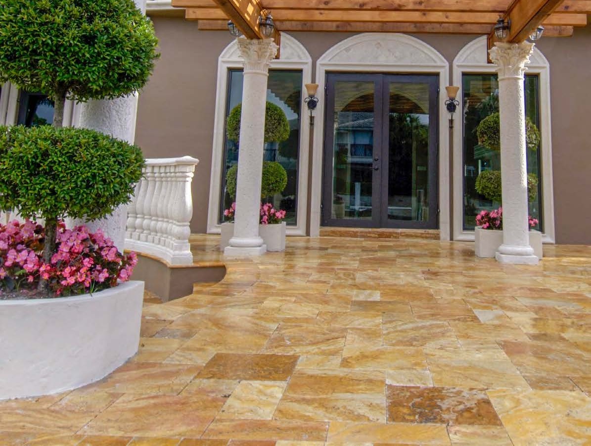 Entrée de villa, dallage travertin doré au premier plan, colonnes et végétation en pots.
